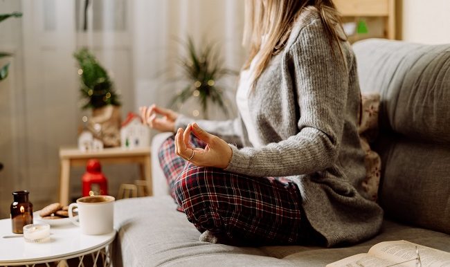 Femme confortable en pull chaud d’hiver tricoté et pyjama à carreaux buvant du chocolat chaud ou du café dans une tasse, livre de lecture, yoga de méditation sur le canapé à la maison.