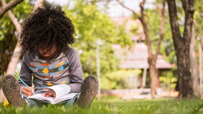 Un petit garçon écrivant sur un cahier assis sur l'herbe verte dans un parc