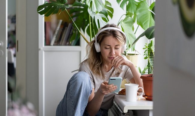 Woman listening to music wear wireless white headphones using mobile smart phone, drinks tea at home